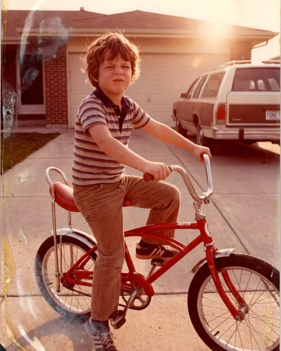 Kinderfoto uit 1982: een jongen op een rode fiets in een oprit, met de avondzon achter hem; de afdruk is naar oranje gezakt en er zitten vlekken in de hoek.