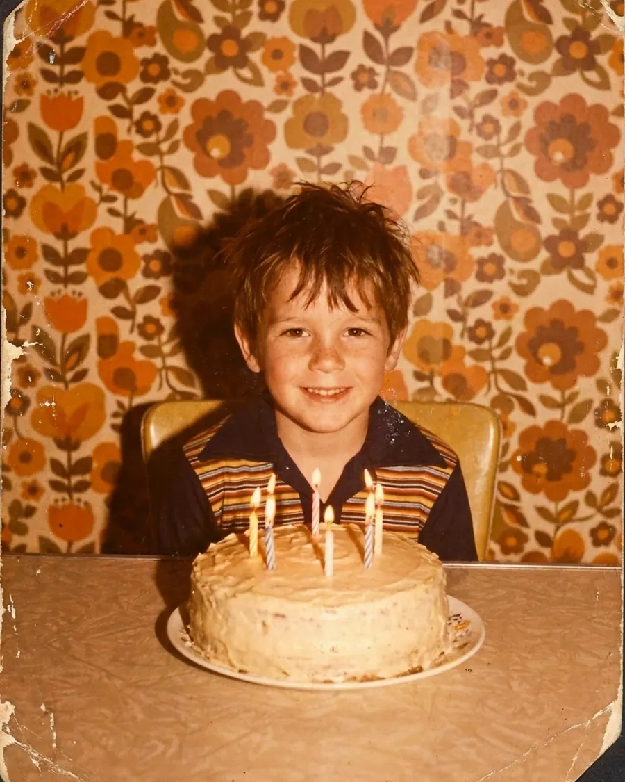 Kinderfoto uit 1972: een jongen achter een verjaardagstaart met vier kaarsjes, voor bloemetjesbehang; de kleuren zijn naar bruin en oranje gekanteld en de randen zijn versleten.