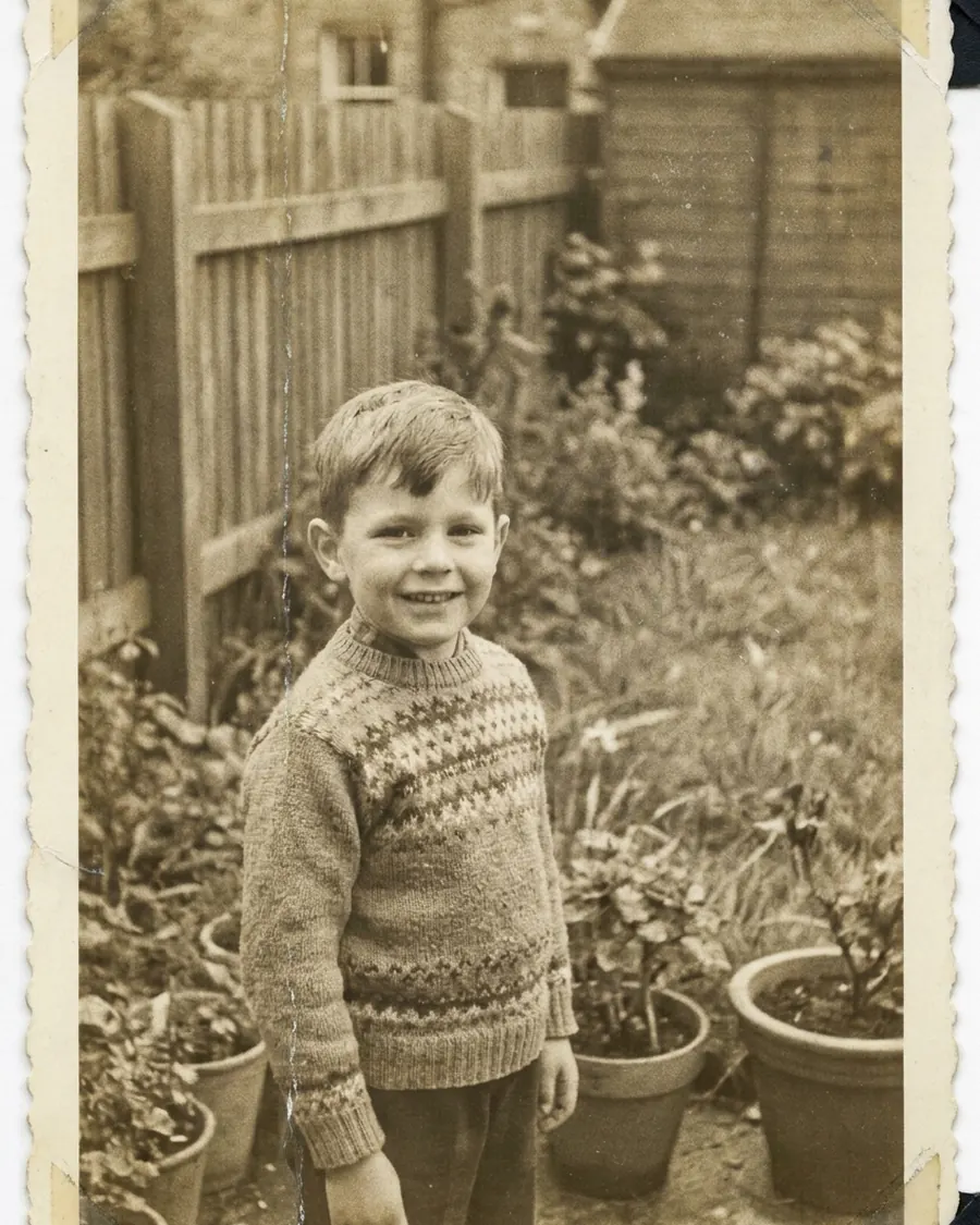 Kinderfoto uit 1962: een jongen in een gebreide trui in een tuin, met een schutting en bloempotten achter hem; de afdruk heeft een sepiawaas en een gekartelde rand.