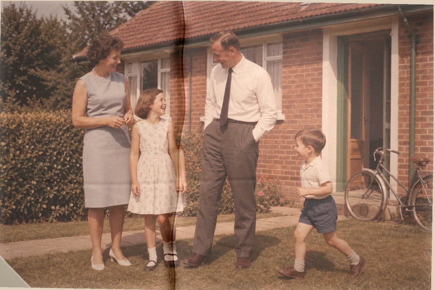 Kleurenfoto uit 1963: een gezin van vier lacht met elkaar in de tuin voor een bakstenen huis, het jongste kind midden in een stap. De afdruk is naar rood gezakt en er loopt een vouw doorheen, met sleet langs de randen.